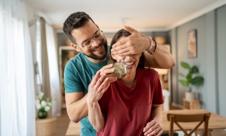 Happy man playfully covering woman's eyes while revealing a glass jar brimming with money, delighting her with their impressive savings at home. Joyful expressions fill the room